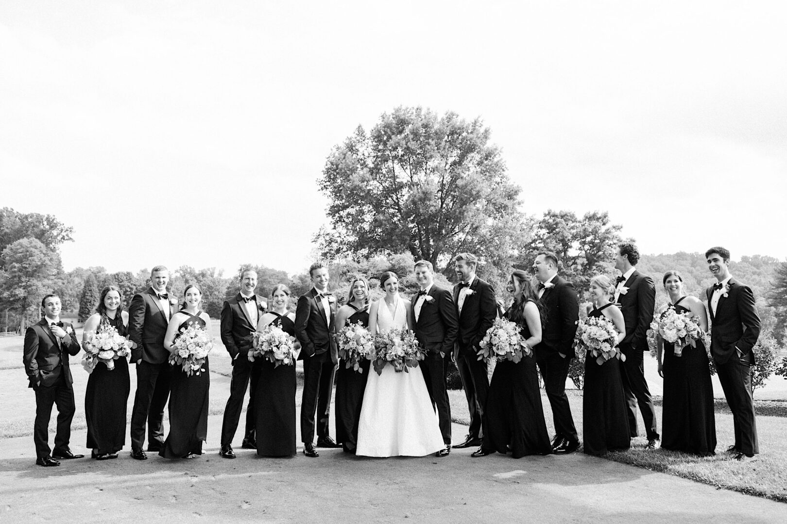 A wedding party poses outdoors in formal attire at the picturesque Overbrook Golf Club. The group consists of bridesmaids in black dresses, groomsmen in black suits, and the bride and groom at the center holding bouquets, capturing a beautiful moment near Philadelphia.