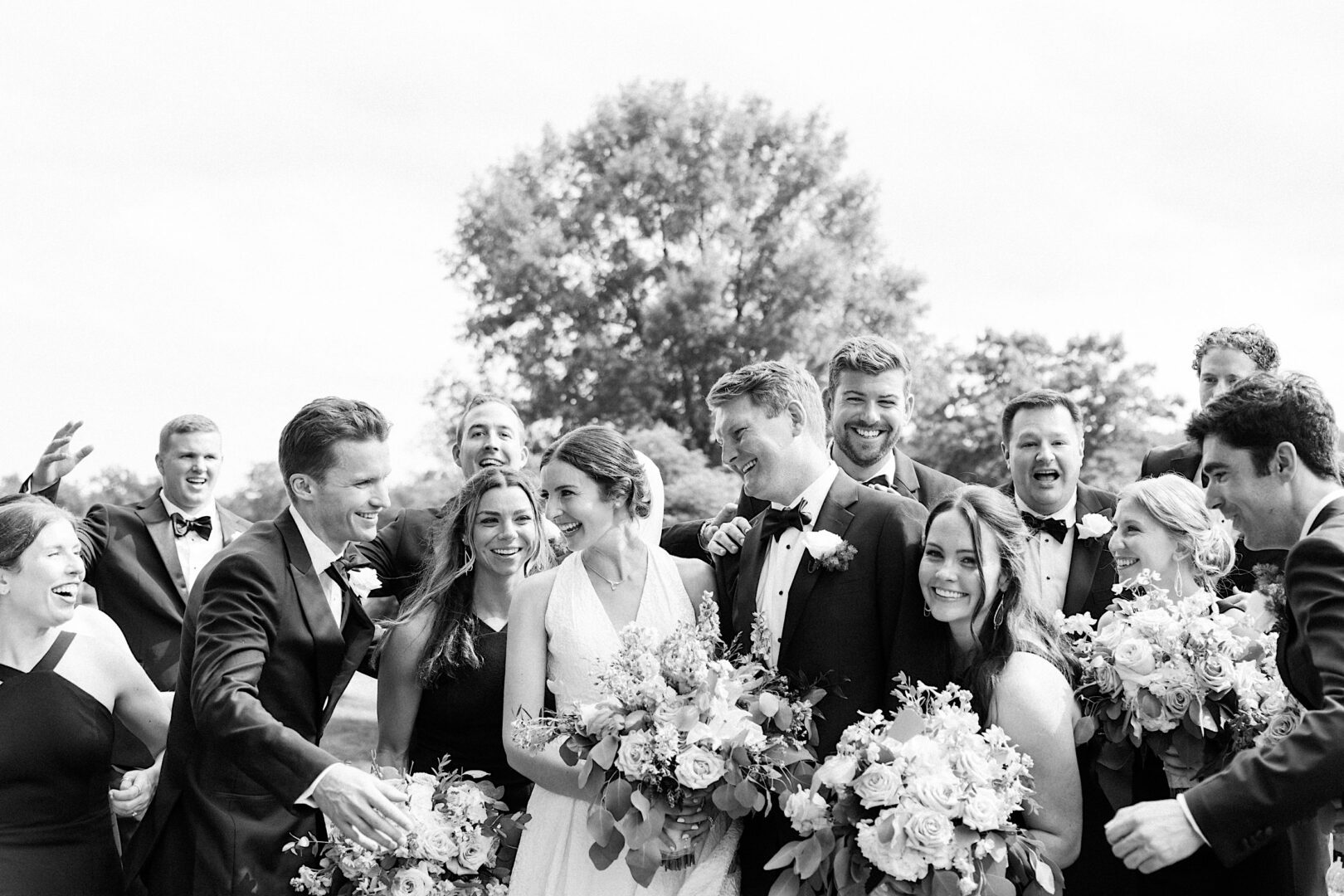 A black and white photo of a group of people dressed formally, celebrating outdoors with bouquets of flowers. The group appears joyful and lively, possibly at an Overbrook Golf Club wedding in Philadelphia. In the background, there are trees and an open sky.