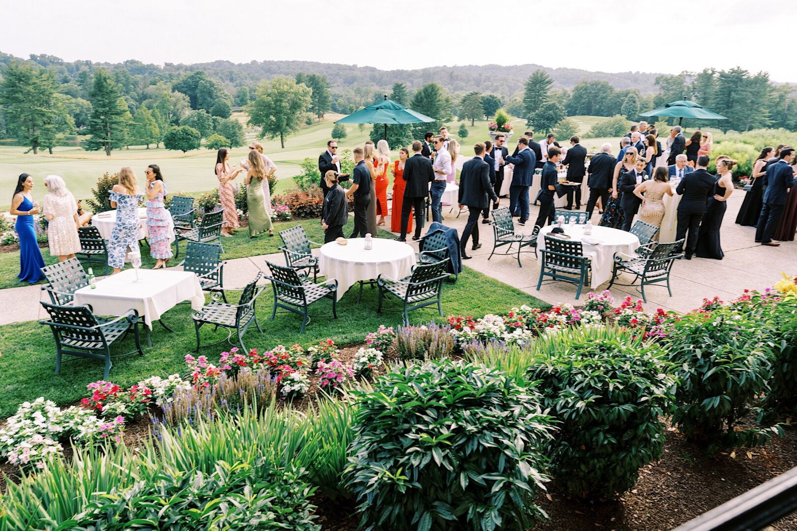 A group of people socializing outdoors at an Overbrook Golf Club wedding, surrounded by tables, chairs, and colorful flowers, with a scenic backdrop of trees and hills.