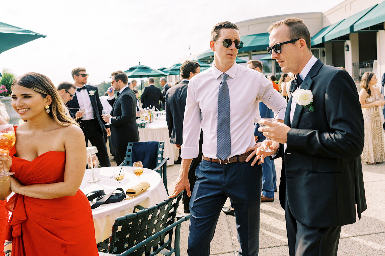 People socializing at an Overbrook Golf Club Wedding, with men in formal attire and sunglasses, and a woman in a red dress holding a drink. Tables with drinks and umbrellas are visible in the background, encapsulating the charm of a Philadelphia event.