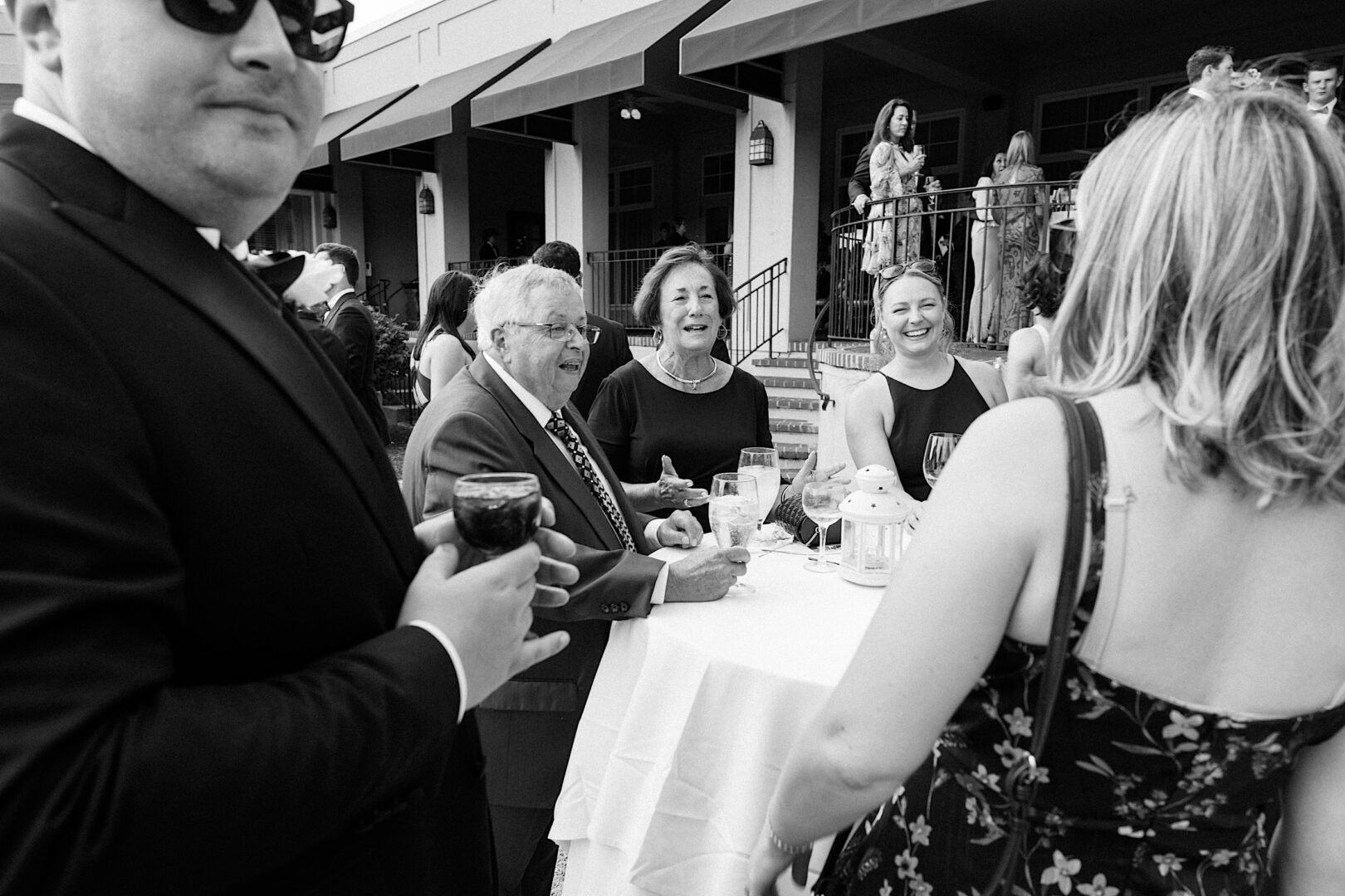 A group of people, dressed formally, stand and converse around a tall table at an outdoor event in Philadelphia. Drinks and a lantern are on the table, setting a charming scene at what appears to be an Overbrook Golf Club wedding near Villanova.