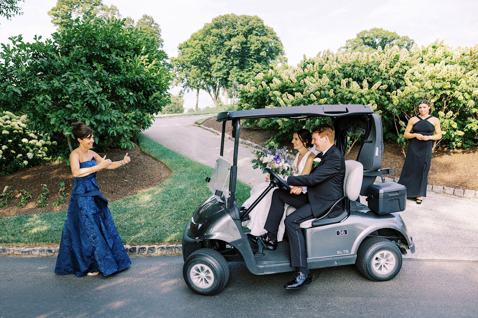 A couple in formal attire rides a golf cart on a paved path at Overbrook Golf Club, while a woman in a blue dress gestures towards them and another woman stands in the background. Greenery surrounds the scene, capturing the essence of their picturesque Philadelphia wedding.