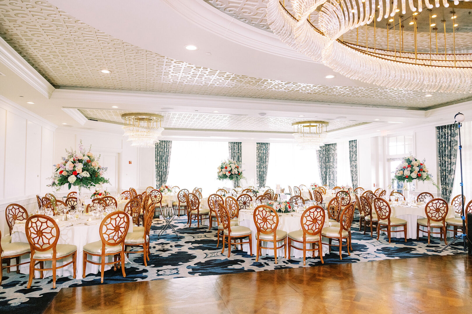 A large banquet hall at Overbrook Golf Club set up for a wedding event, featuring round tables with floral centerpieces, wooden chairs with intricate backs, decorative ceiling, chandeliers, and patterned carpet. Located in Villanova near Philadelphia.