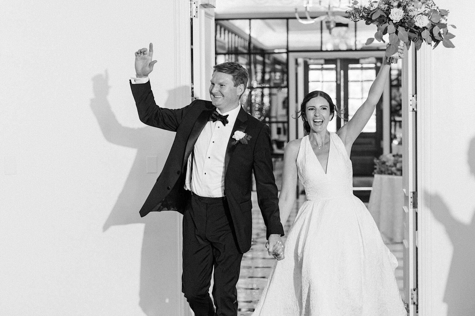 A couple, dressed in formal wedding attire, joyfully enters a room holding hands at their Overbrook Golf Club Wedding with the woman raising a bouquet and both smiling. The scene, set in Villanova near Philadelphia, is captured beautifully in black and white.