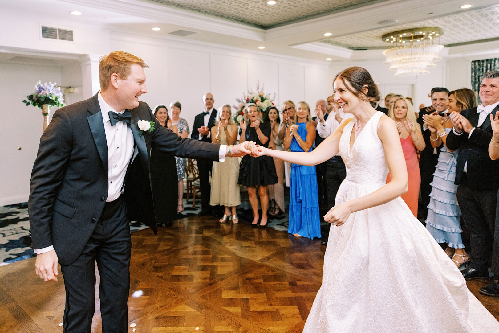 A couple dances together on a polished wooden floor, dressed in formal attire, surrounded by applauding guests in an elegant room at an Overbrook Golf Club wedding near Philadelphia.