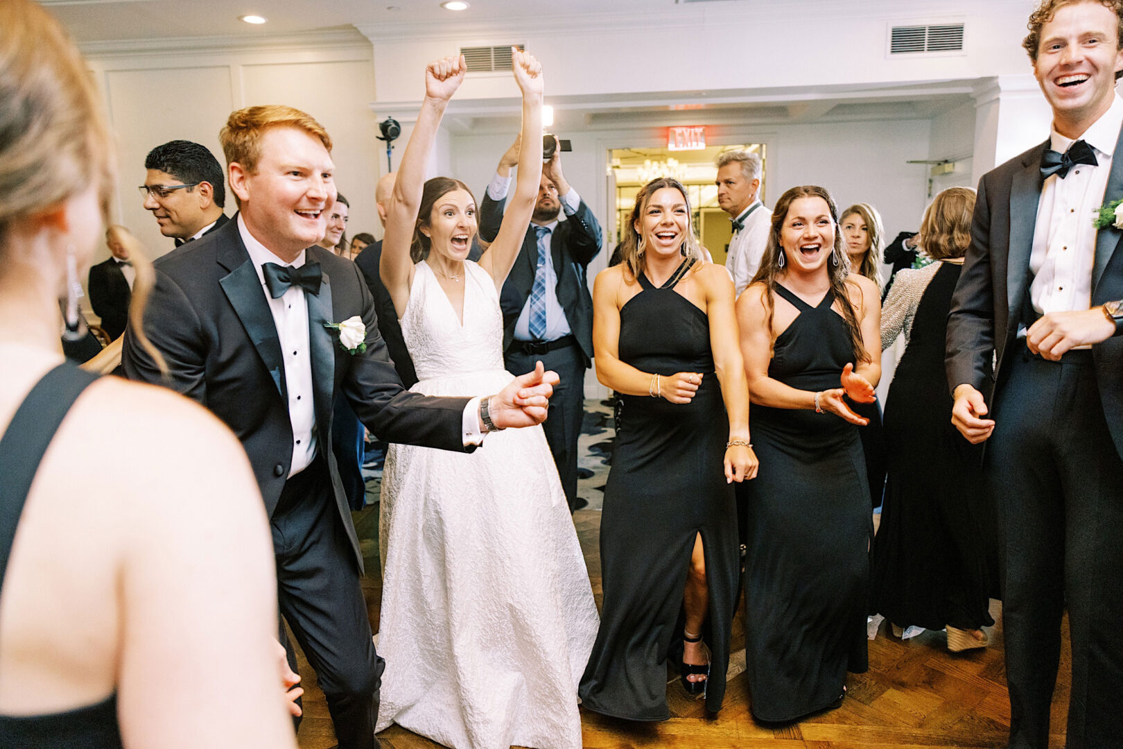 A group of people, including a bride and groom, dressed in formal attire, celebrate joyfully indoors at an Overbrook Golf Club wedding. The bride has her arms raised and guests around her are smiling and cheering at this beautiful Villanova event.