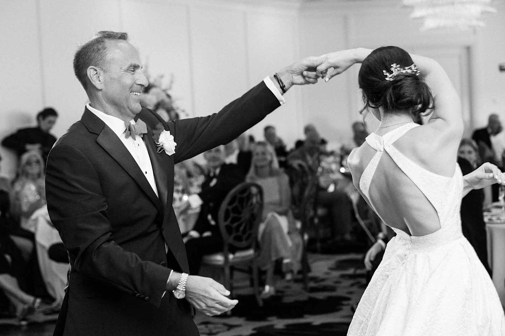 A man and a woman, both dressed formally, dance together at an Overbrook Golf Club Wedding in Philadelphia. The woman is wearing a sleeveless dress with a cross-back design. The setting appears to be a celebration or event filled with seated guests.