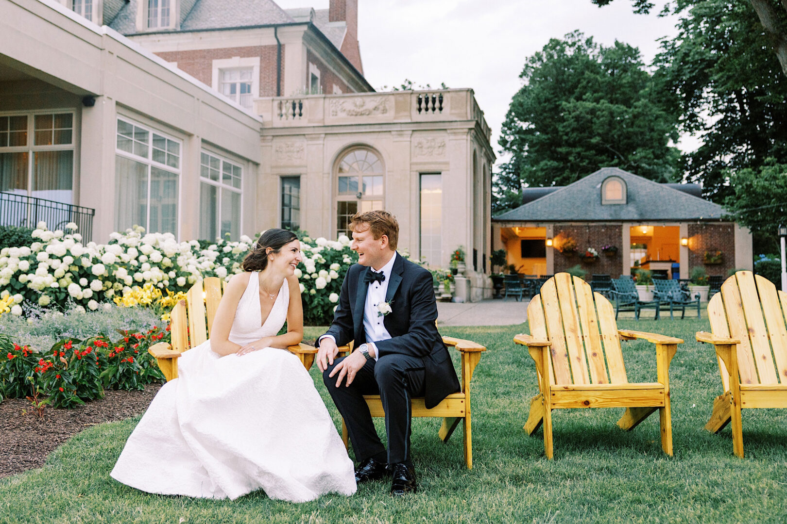 A bride and groom sit on yellow wooden chairs, smiling at each other. They are outdoors in front of a large building with flowers and trees in the background, celebrating their beautiful Overbrook Golf Club wedding in Villanova.