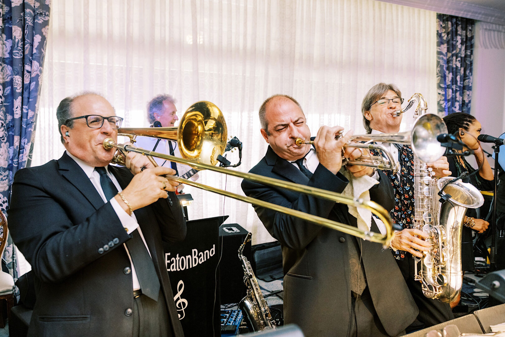 A band of musicians in suits plays various brass instruments, including trombones, a trumpet, and a saxophone, in front of a curtain backdrop. A "Band" sign is visible in the background, setting the perfect tone for an elegant Overbrook Golf Club Wedding near Villanova in Philadelphia.