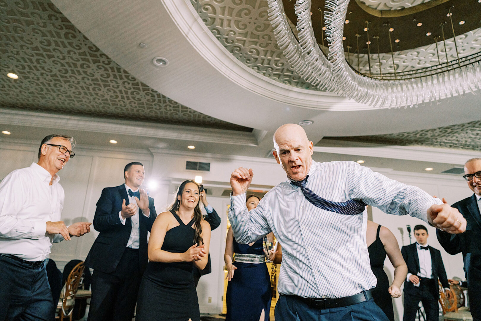 At the Overbrook Golf Club Wedding in Philadelphia, a group of people in formal attire dance energetically in a banquet hall adorned with a decorative ceiling and a large chandelier.