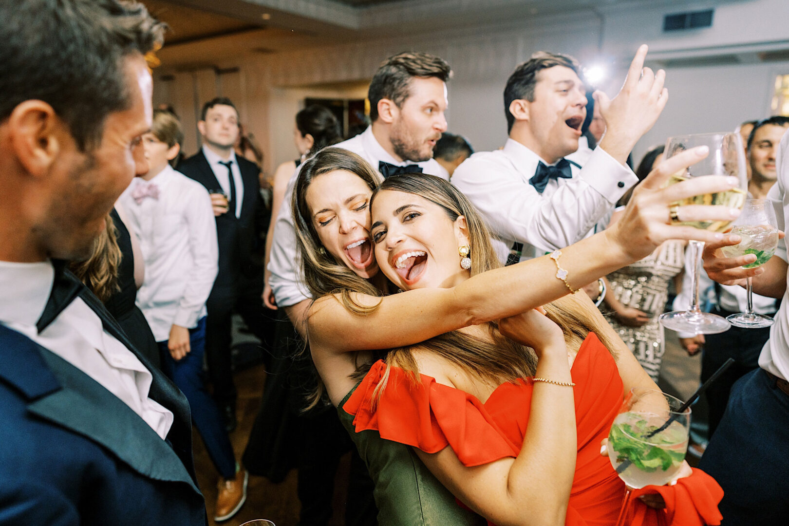 Two women smiling and hugging while taking a selfie at a lively, crowded party; people around them are dancing and celebrating in formal attire. The joyous event took place at an elegant Overbrook Golf Club wedding near Philadelphia.