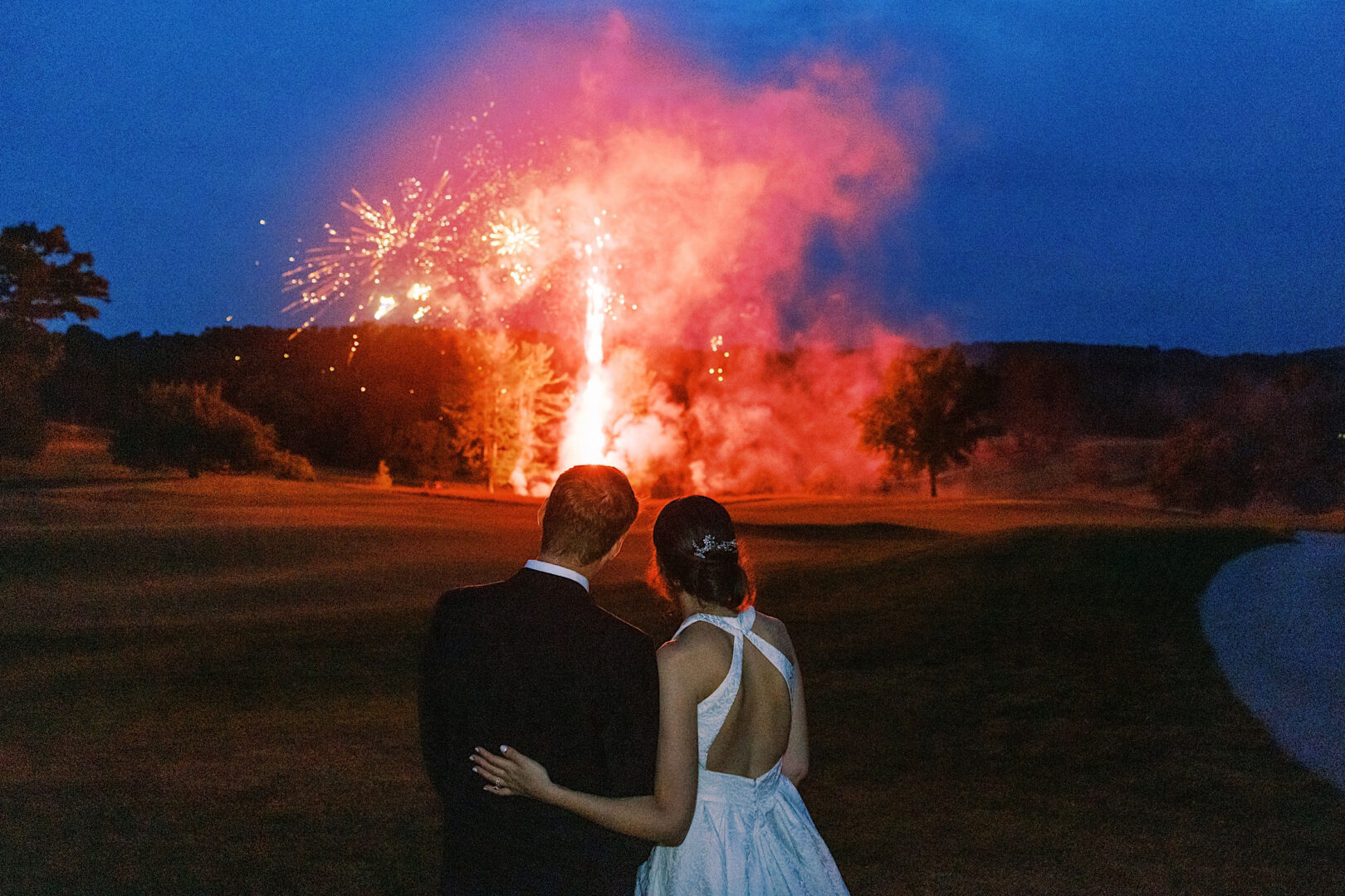 A couple dressed in formal attire watches a bright fireworks display in the night sky over a grassy field at an enchanting Overbrook Golf Club wedding.