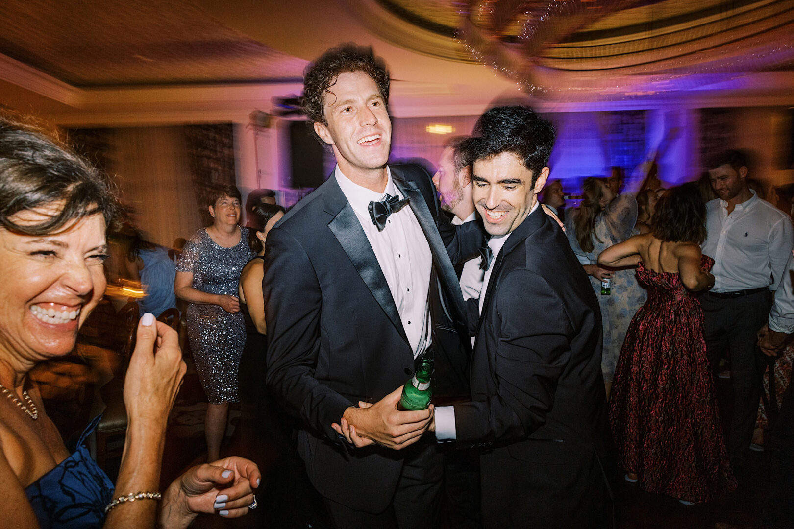 People in formal attire are dancing and laughing at a party. Two men in tuxedos are in the center, one holding a bottle, surrounded by joyful guests. The lively event at Overbrook Golf Club Wedding near Villanova captures the celebratory essence of Philadelphia's vibrant social scene.