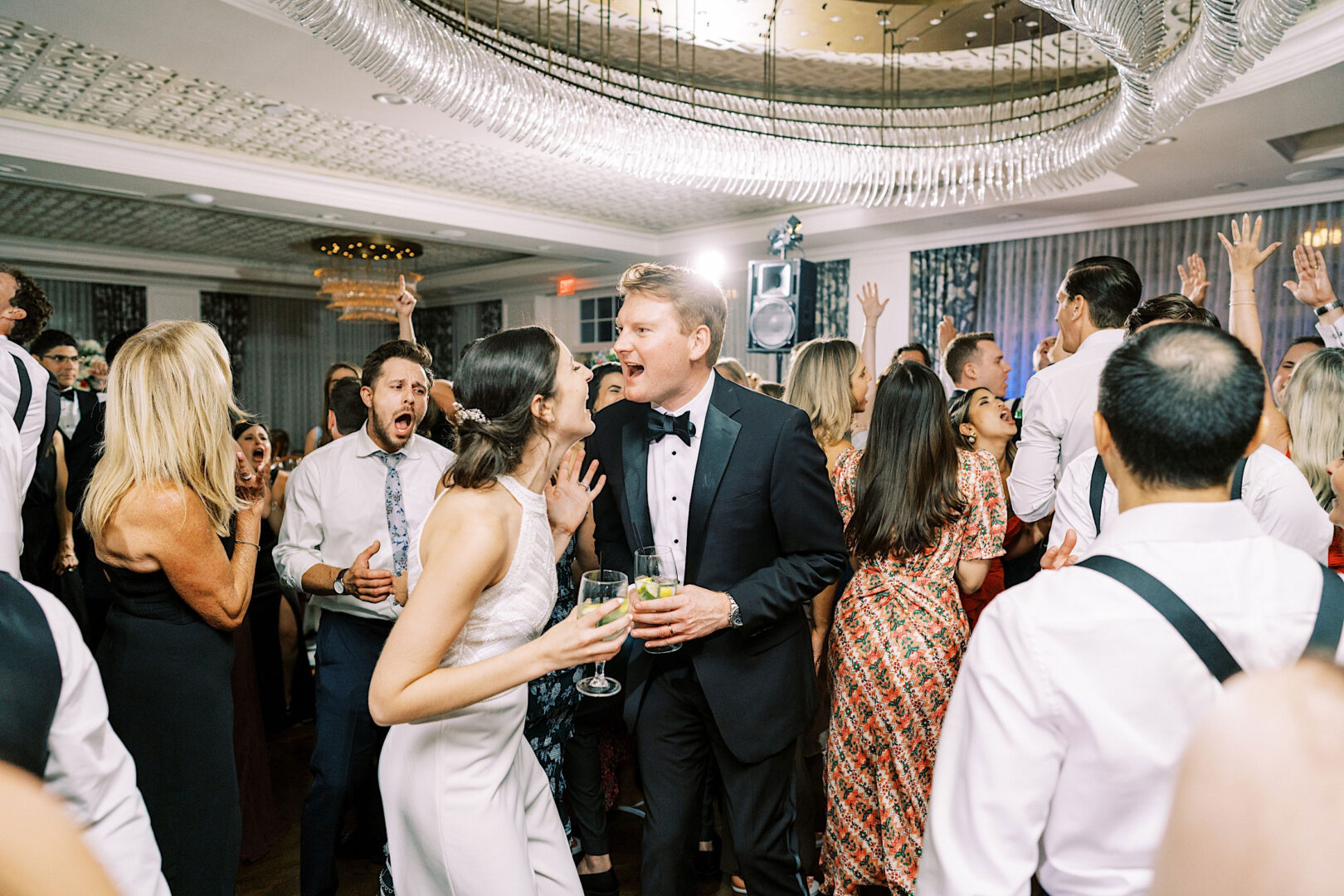 At an Overbrook Golf Club Wedding in Villanova, a bride and groom, dressed in formal attire, sing together surrounded by guests dancing at a lively wedding reception.