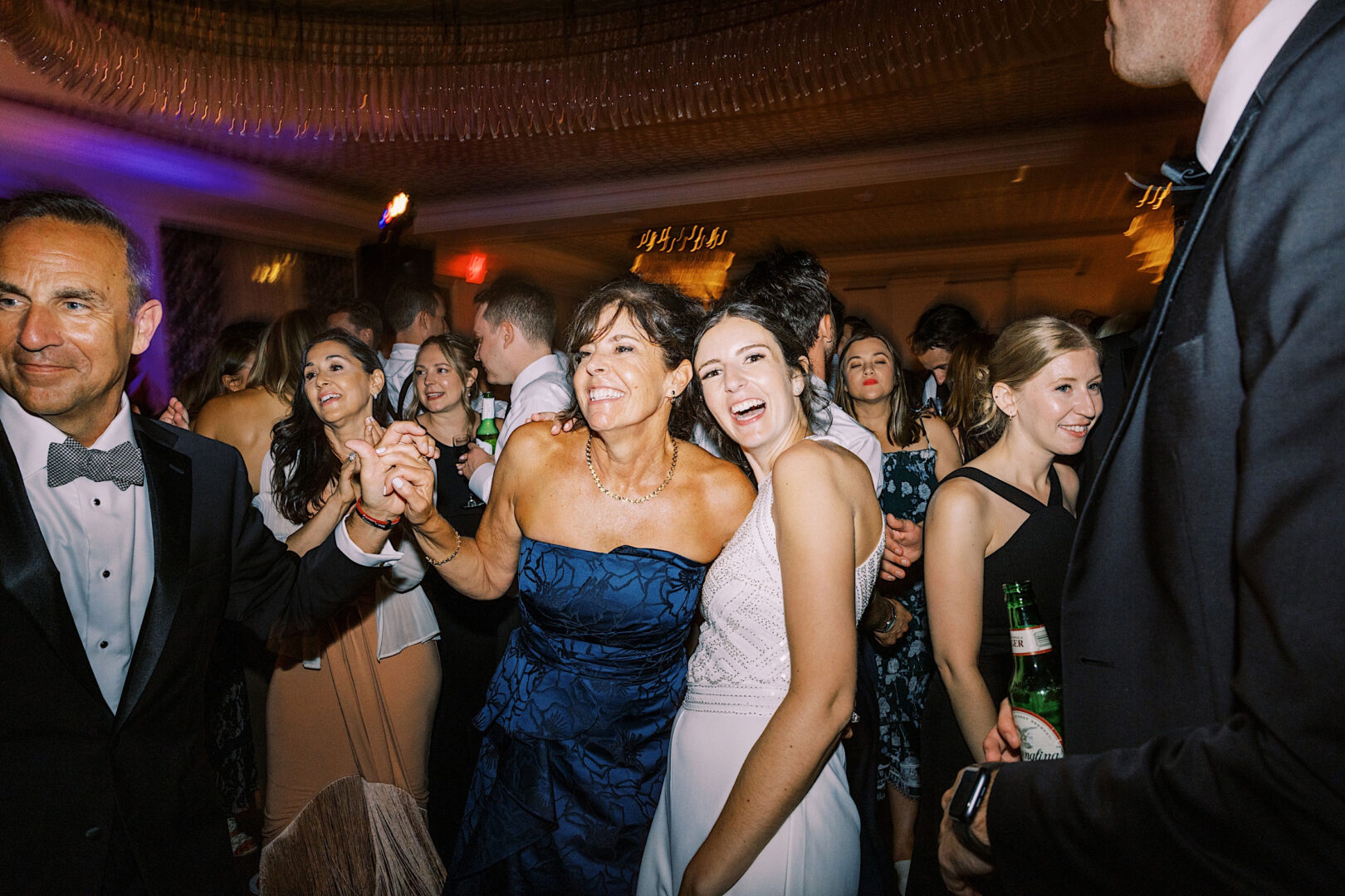 A group of people in formal attire are celebrating at an indoor event. At the Overbrook Golf Club Wedding near Villanova, a woman in a blue dress and a woman in a white dress are smiling and holding hands in the foreground.