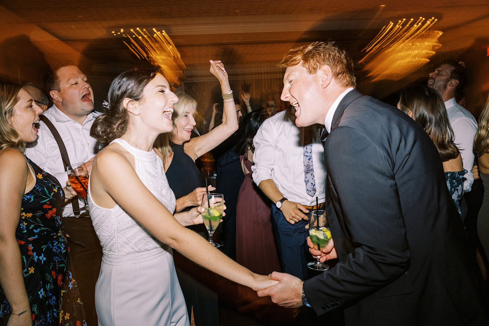 At their beautiful Overbrook Golf Club wedding in Villanova, a bride and groom joyfully dance while holding hands and drinks, surrounded by cheering guests in a lively reception setting.