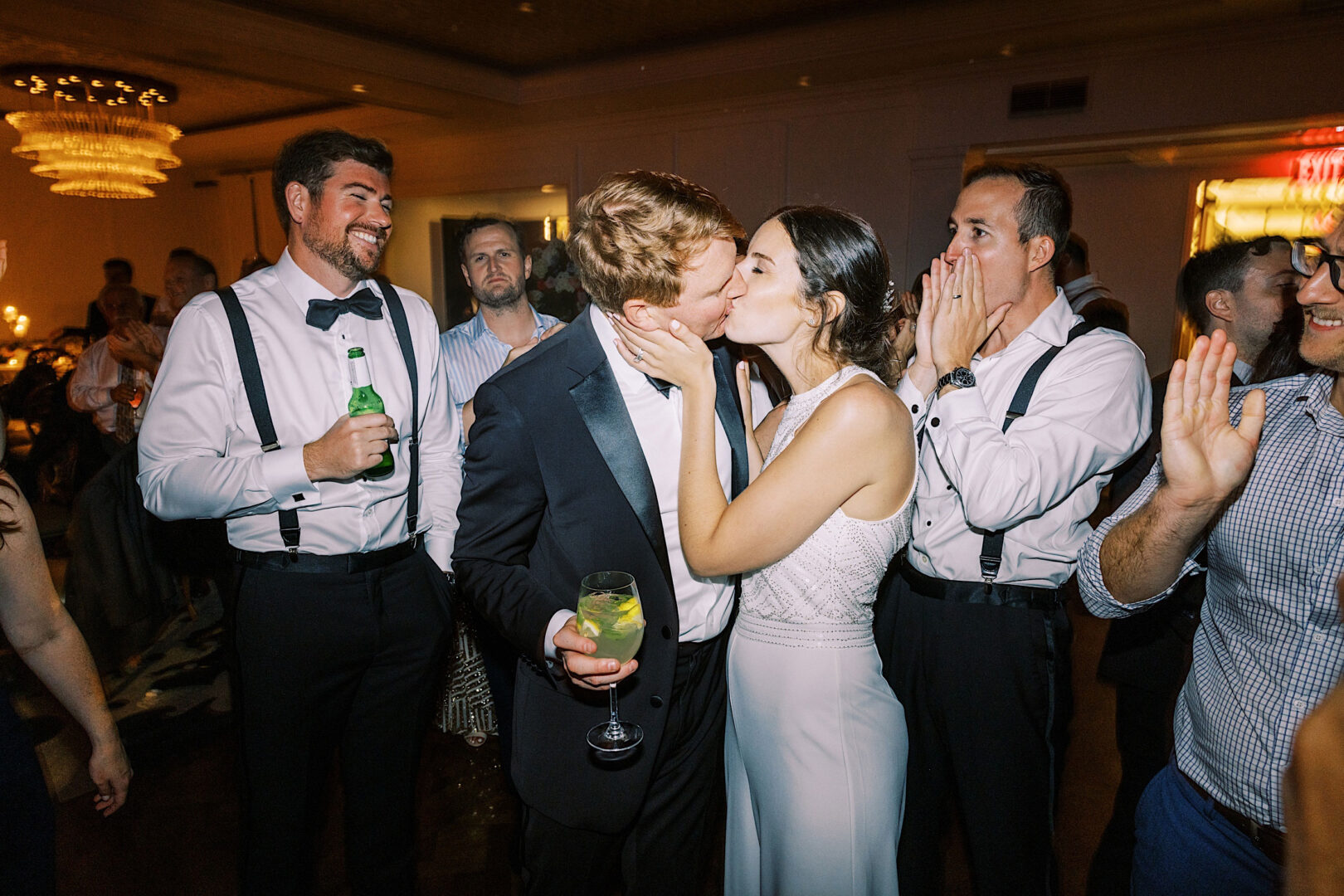 At an Overbrook Golf Club Wedding in Villanova, a couple kisses at a celebration while guests around them cheer. The man on the left is holding a beer, and the person on the right appears to be clapping.