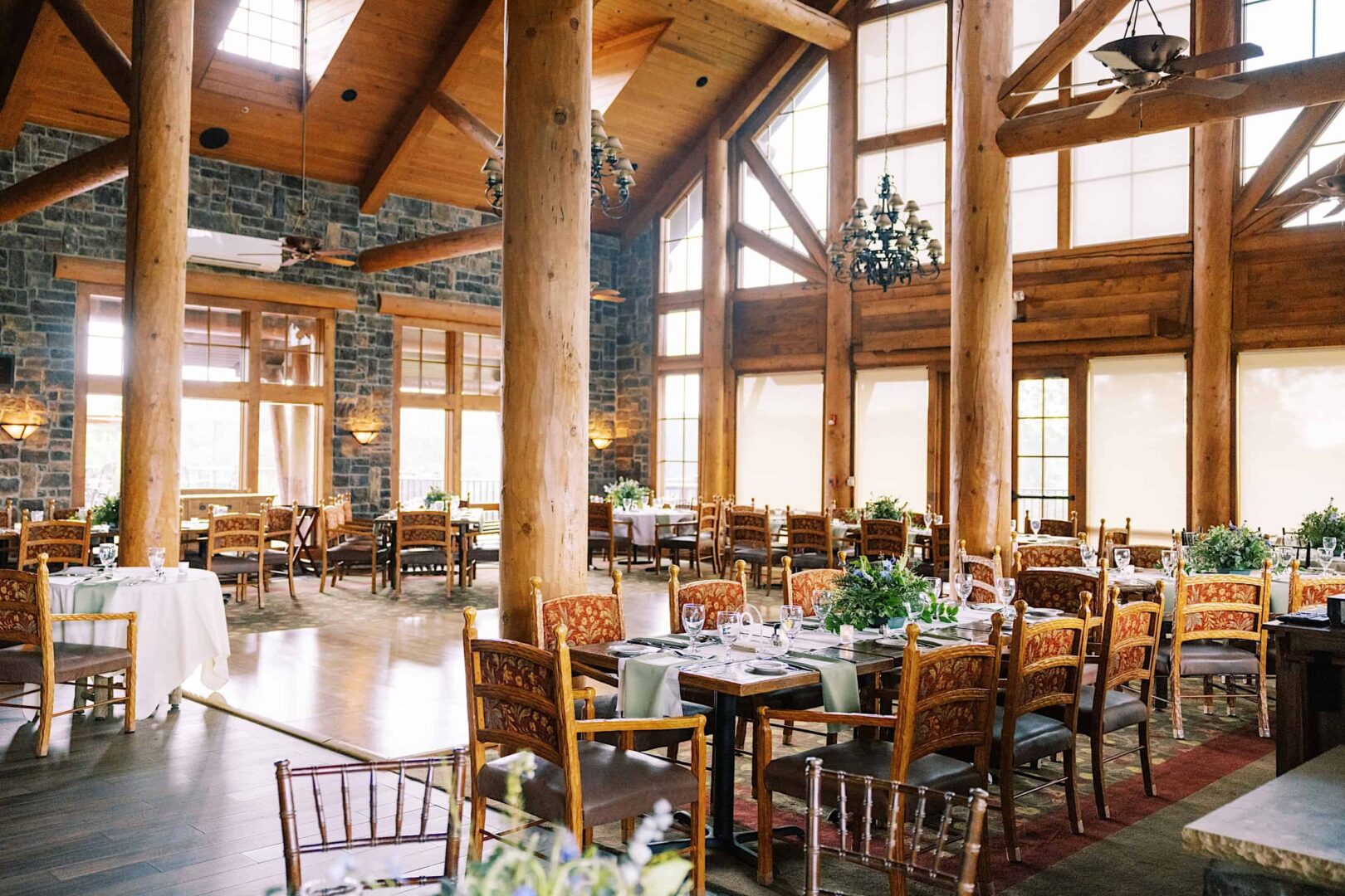 A spacious dining area with wooden beams, stone walls, and large windows at a charming New Jersey country club. Tables are set with white linens and adorned with green centerpieces, surrounded by wooden chairs with cushioned seats—perfect for an elegant wedding reception.