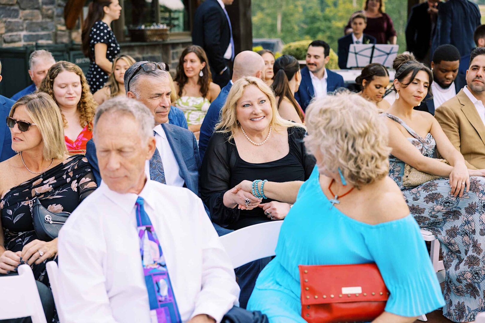 A group of people, dressed formally, sit on white chairs at an outdoor event at The Ridge at Back Brook Country Club in New Jersey. A woman in blue and a woman in black are shaking hands in the center. Trees and other attendees are in the background.