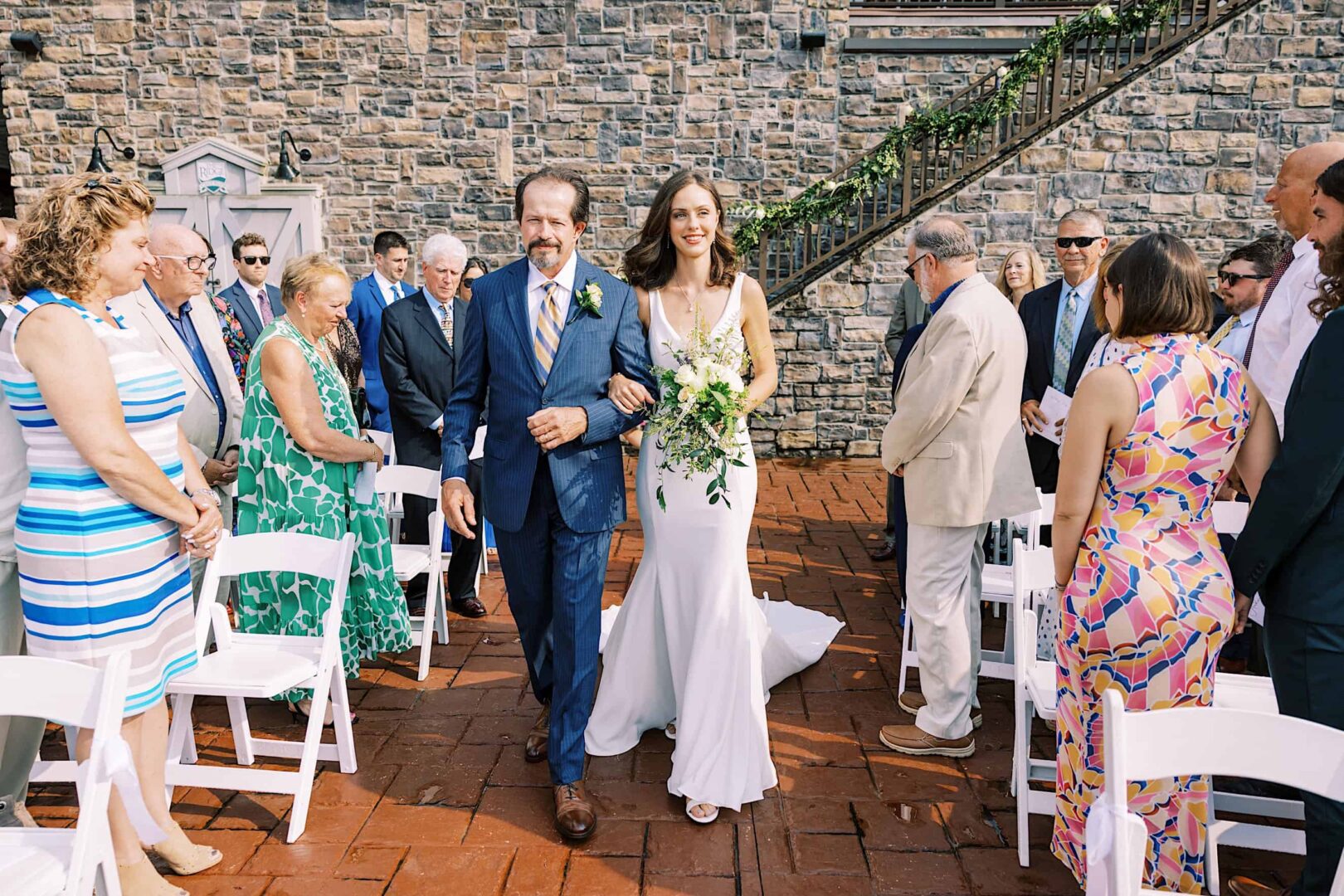 A bride in a white gown walks down the aisle with an older man in a suit at an outdoor wedding at The Ridge at Back Brook, New Jersey, surrounded by seated guests.