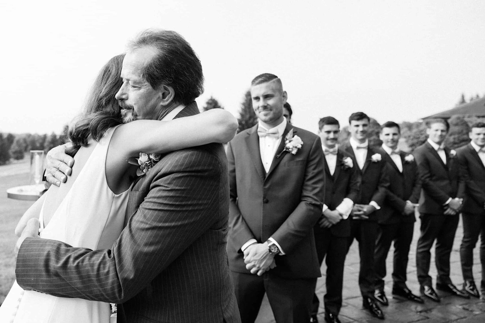 A bride embraces an older gentleman in a suit, while a smiling groom and groomsmen stand in a line behind them. The scene appears to be outdoors at The Ridge at Back Brook, capturing the joy of a beautiful New Jersey wedding.