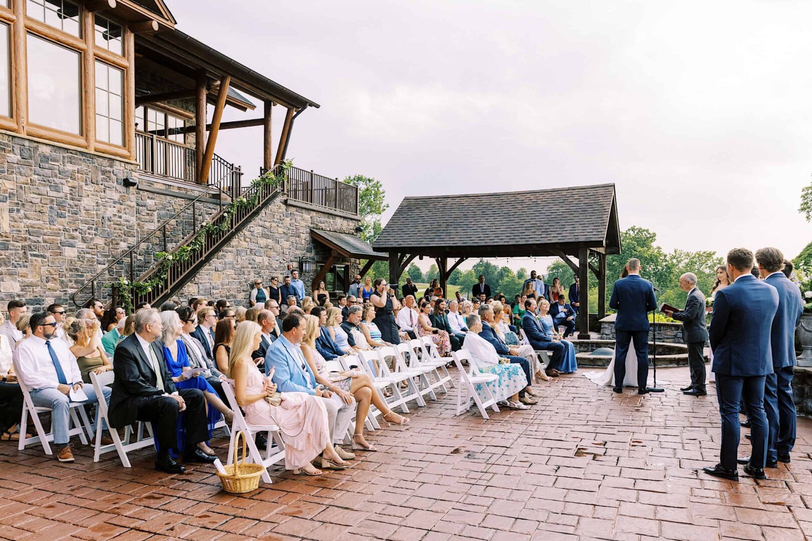 A wedding ceremony takes place outdoors at The Ridge at Back Brook, with guests seated on white chairs. The event is held near a stone building with a wooden pergola, and people are dressed in formal attire. This picturesque New Jersey country club setting adds an extra touch of elegance to the occasion.