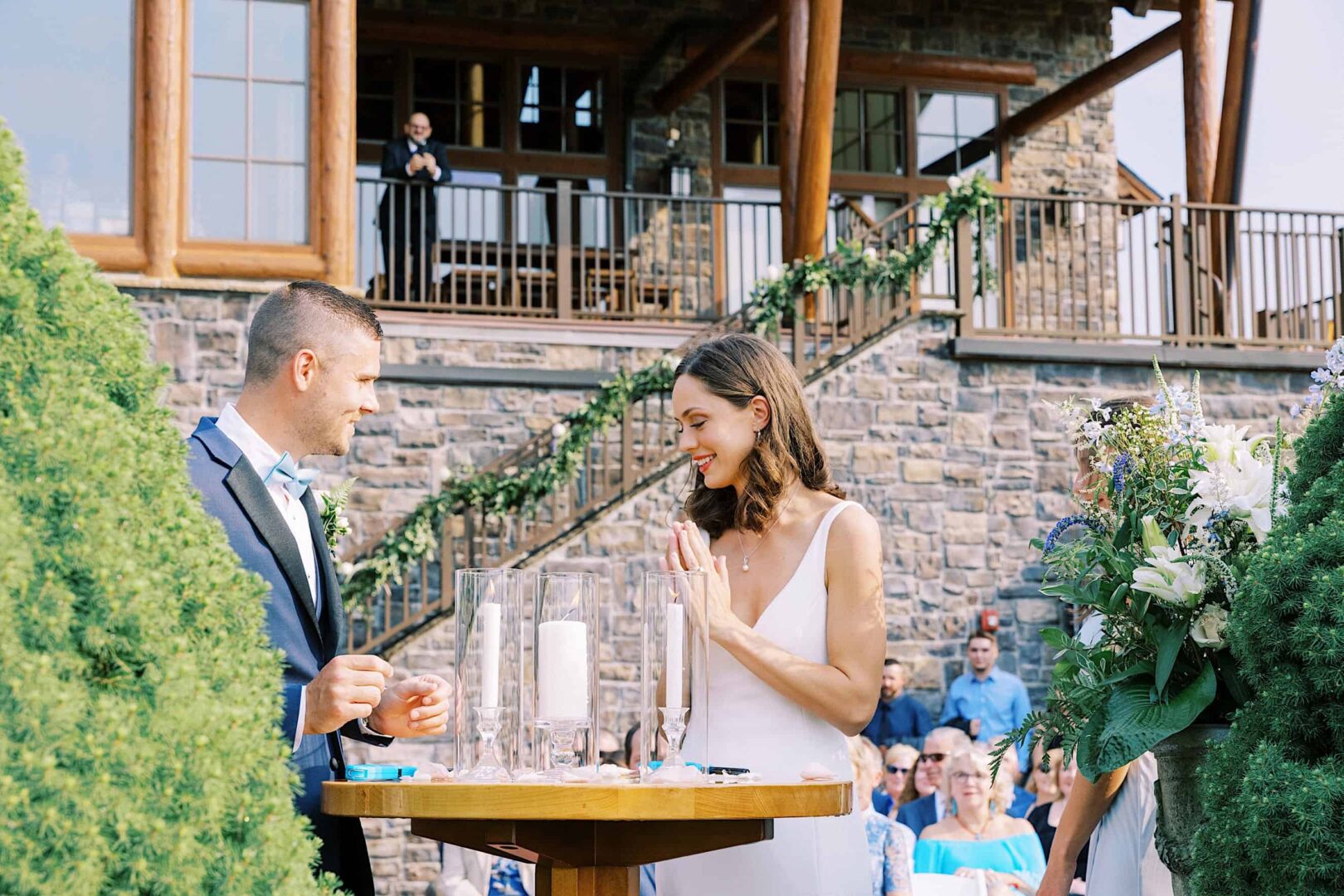 A couple stands at a round table during an outdoor wedding ceremony in New Jersey. Both are looking at each other, wearing formal attire, with guests seated in the background and a stone building behind them, capturing the elegance of their special day at the country club.