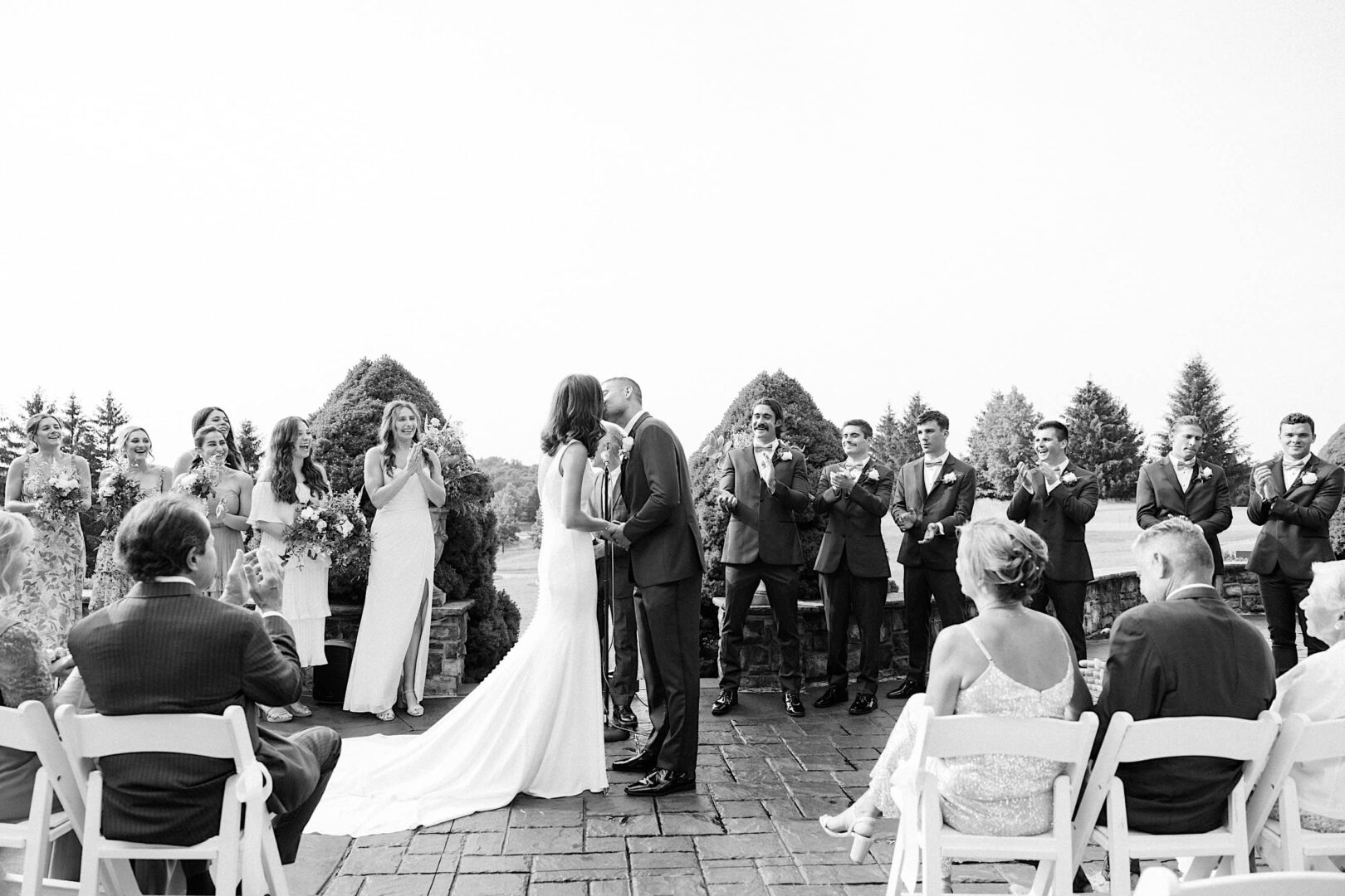 At The Ridge at Back Brook, a bride and groom share a kiss during their outdoor wedding ceremony. Surrounded by their bridal party, guests seated in white chairs watch and clap, creating an unforgettable moment at this beautiful country club.