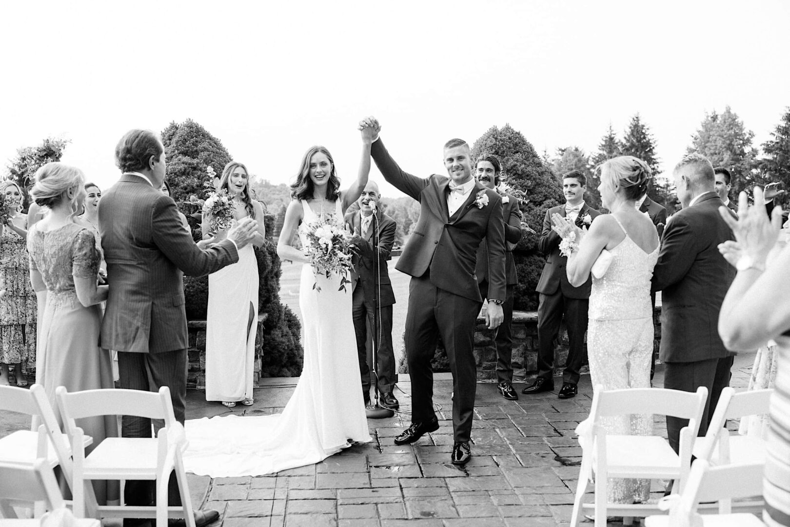 A bride and groom raise their hands in celebration at The Ridge at Back Brook, a New Jersey country club, as guests clap around them during an outdoor wedding ceremony.