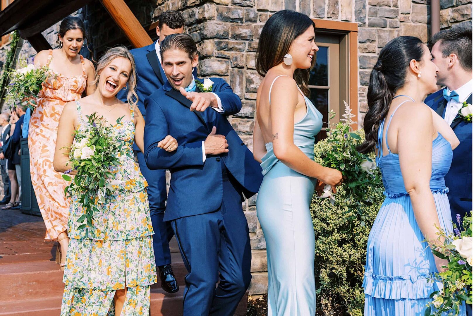 A group of people in formal attire, including floral dresses and blue suits, descend the outdoor steps of The Ridge at Back Brook Country Club, smiling and interacting cheerfully outside a stone building.