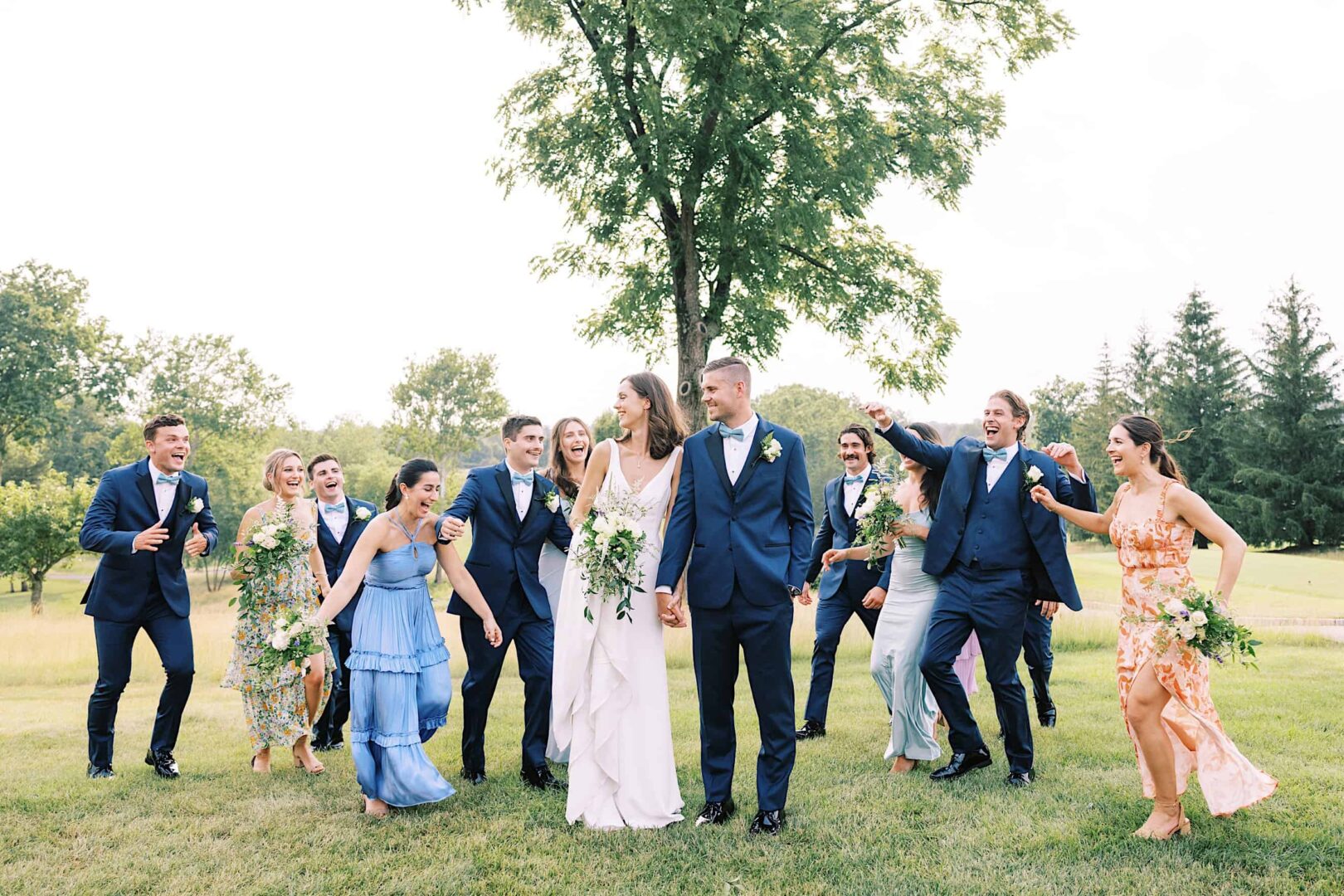 A bride and groom smile and hold hands while being surrounded by a joyful bridal party outdoors at The Ridge at Back Brook in New Jersey.