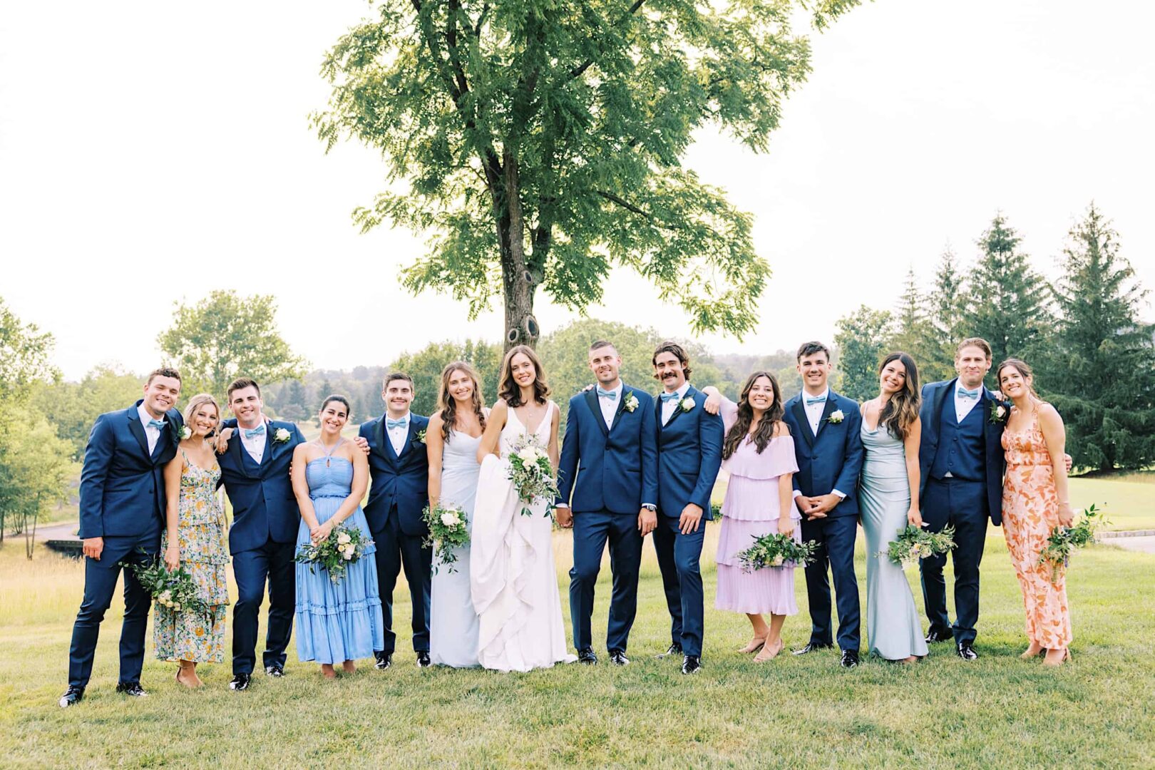 A wedding party stands outdoors at a New Jersey country club; six men in blue suits and six women in assorted dresses pose together, holding bouquets. Trees and greenery are visible in the background.