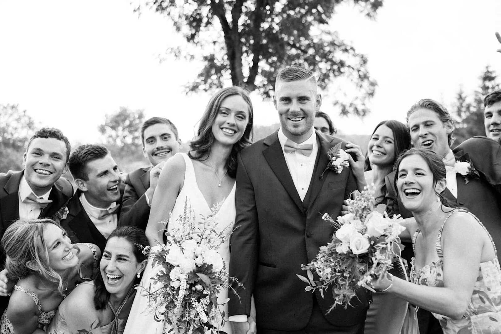 A black-and-white photo of a bride and groom in the center, smiling with a joyfully gathered wedding party around them, holding floral arrangements at The Ridge at Back Brook in New Jersey.