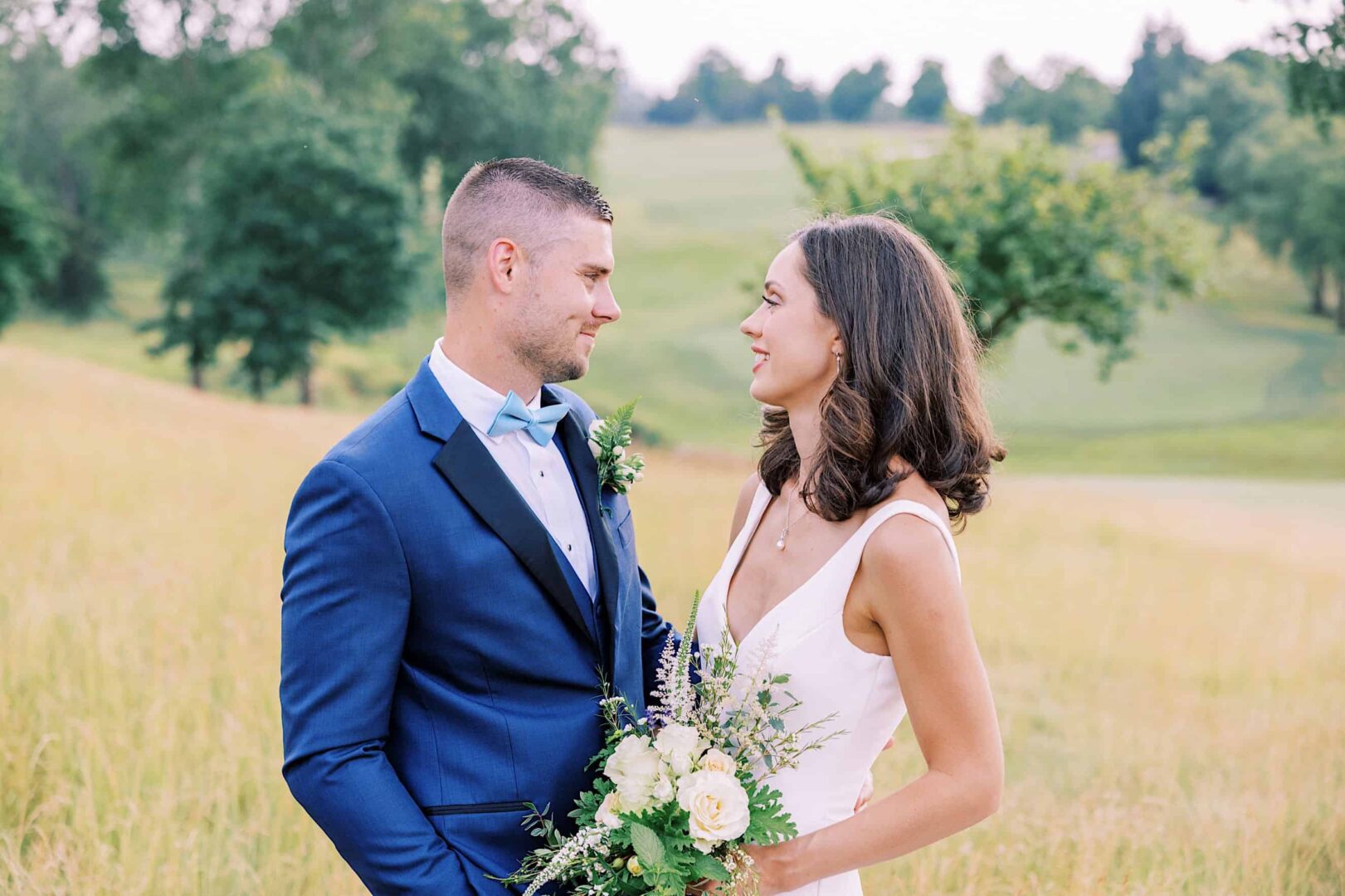 A couple dressed in formal attire are standing in a grassy field, facing each other and smiling. The woman holds a bouquet of flowers, and the landscape features trees and an open field at The Ridge at Back Brook, perfect for a wedding.