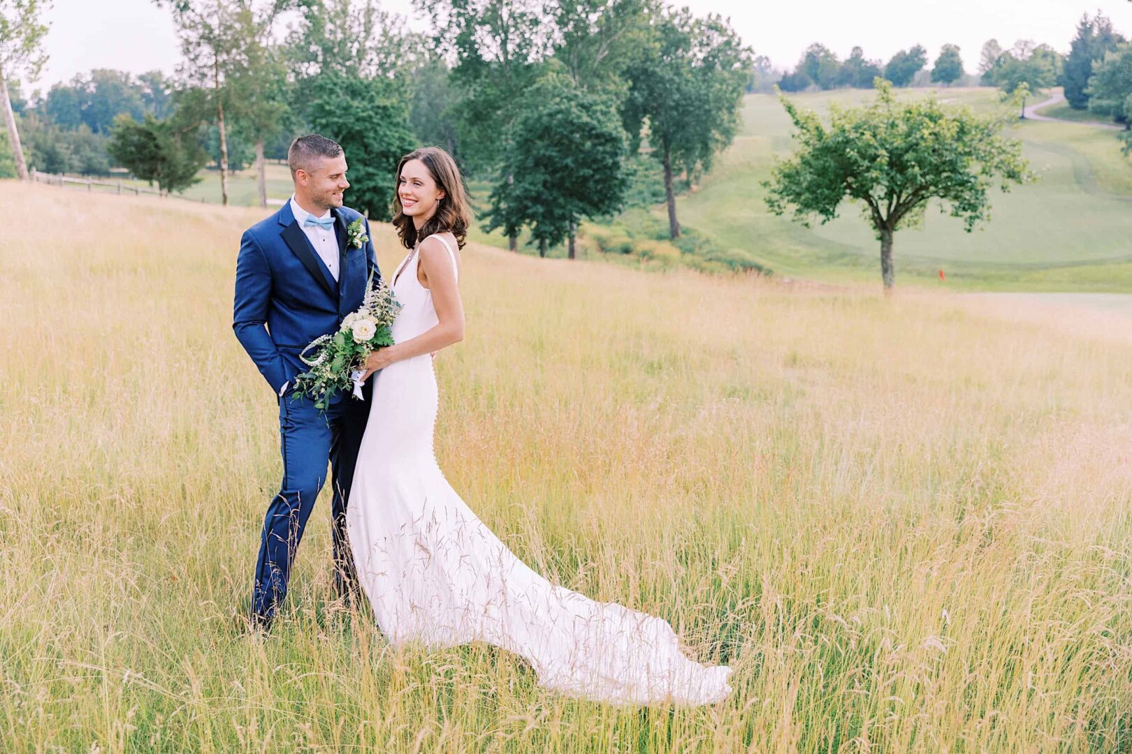 A bride and groom stand in a grassy field at The Ridge at Back Brook, with trees and a golf course in the background. The bride is holding a bouquet and wearing a long white dress, while the groom wears a blue suit. This picturesque New Jersey country club provides an idyllic setting for their special day.