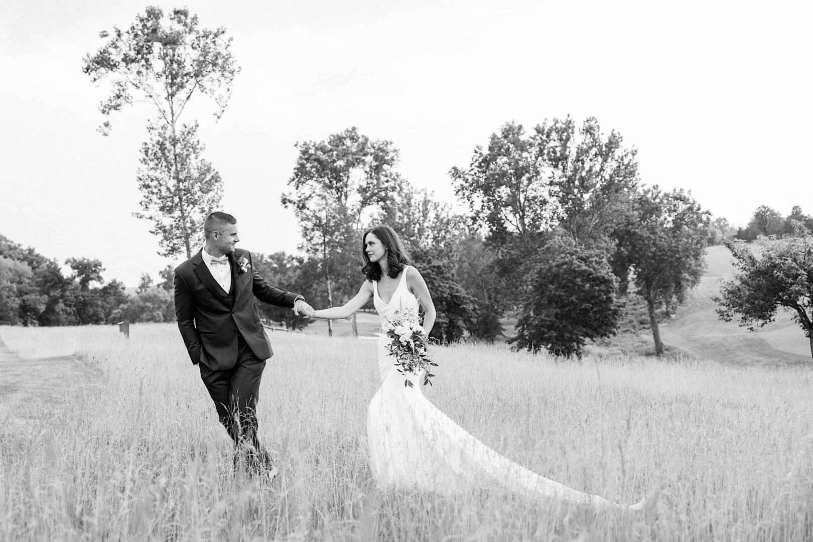 A couple in formal wedding attire walks hand-in-hand through a field at The Ridge at Back Brook. The bride wears a long flowing dress and holds a bouquet, while the groom sports a suit with a boutonniere, creating an enchanting country club wedding scene.