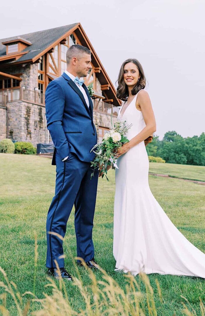 A bride in a white dress and a groom in a blue suit stand on the grassy lawn of The Ridge at Back Brook Country Club in front of a stone building. The bride holds a bouquet while the groom faces her and smiles.