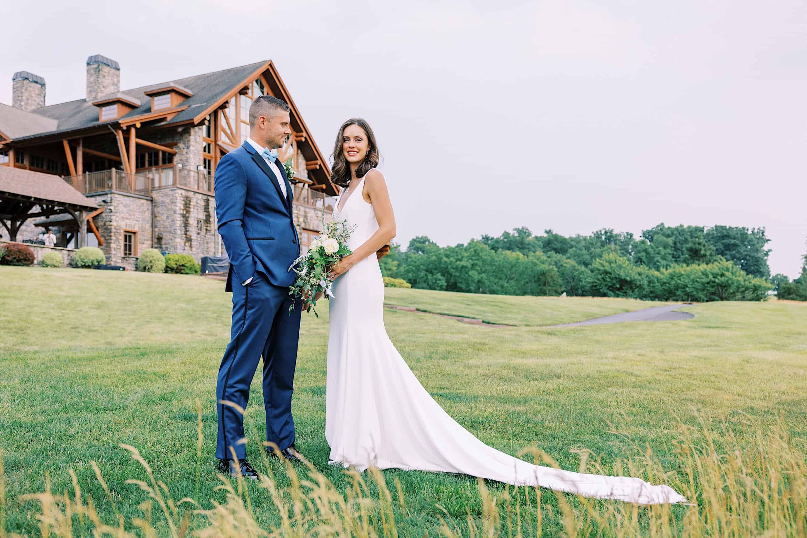 A bride in a white dress and a groom in a blue suit stand on the grassy lawn of The Ridge at Back Brook Country Club in front of a stone building. The bride holds a bouquet while the groom faces her and smiles.