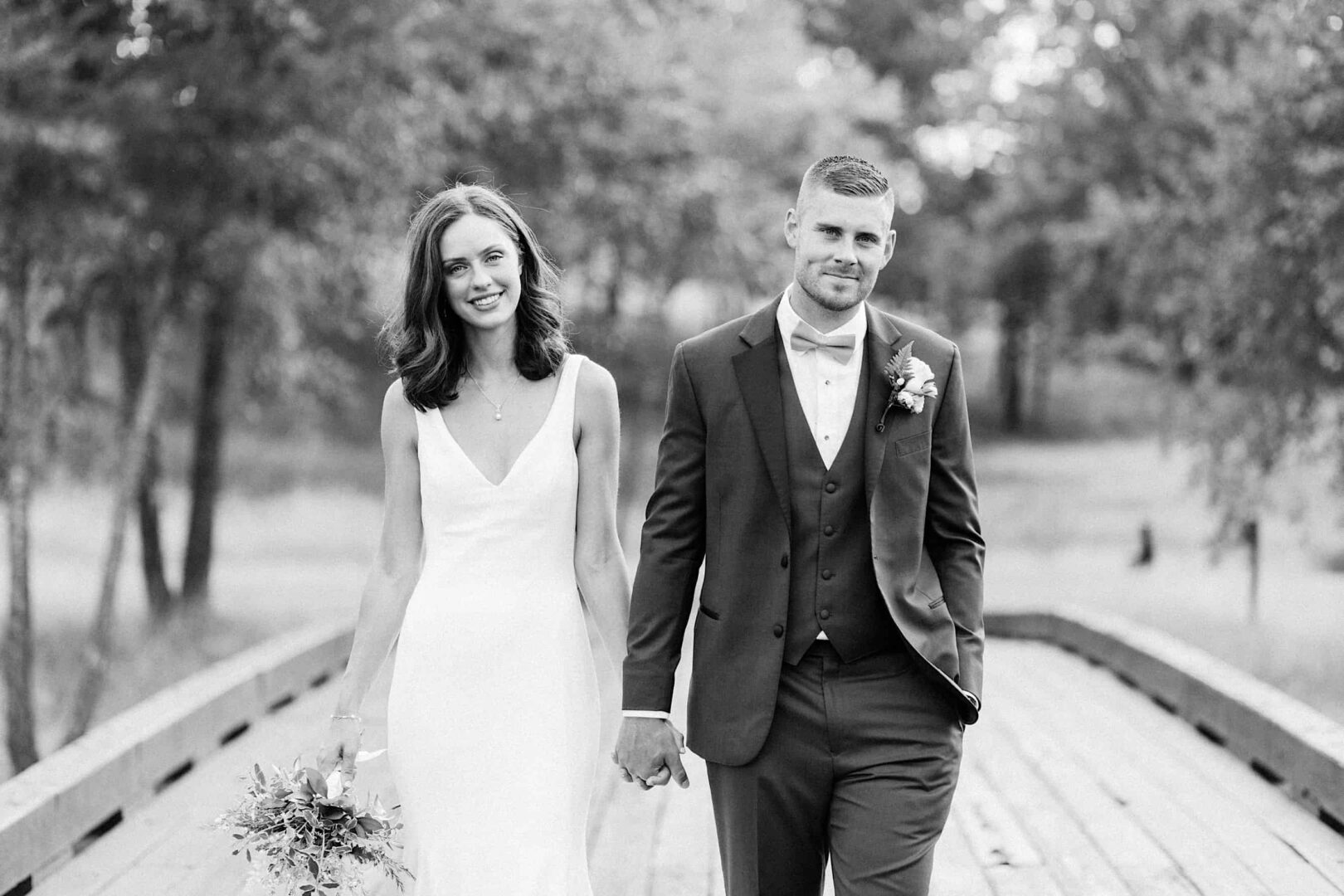 A bride and groom walk hand in hand down a wooden path outdoors at The Ridge at Back Brook, with the bride holding a bouquet and the groom in a suit and bow tie.