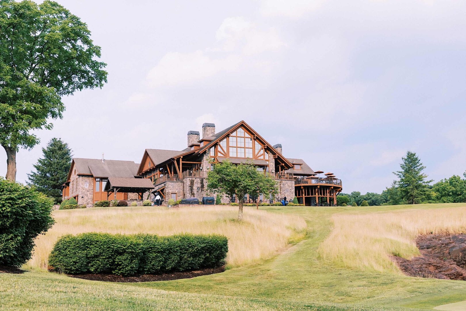 A large, stone and wood lodge with multiple chimneys stands amidst a lush, green landscape under a partly cloudy sky at The Ridge at Back Brook in New Jersey, surrounded by trees and tall grass—a perfect setting for a wedding.