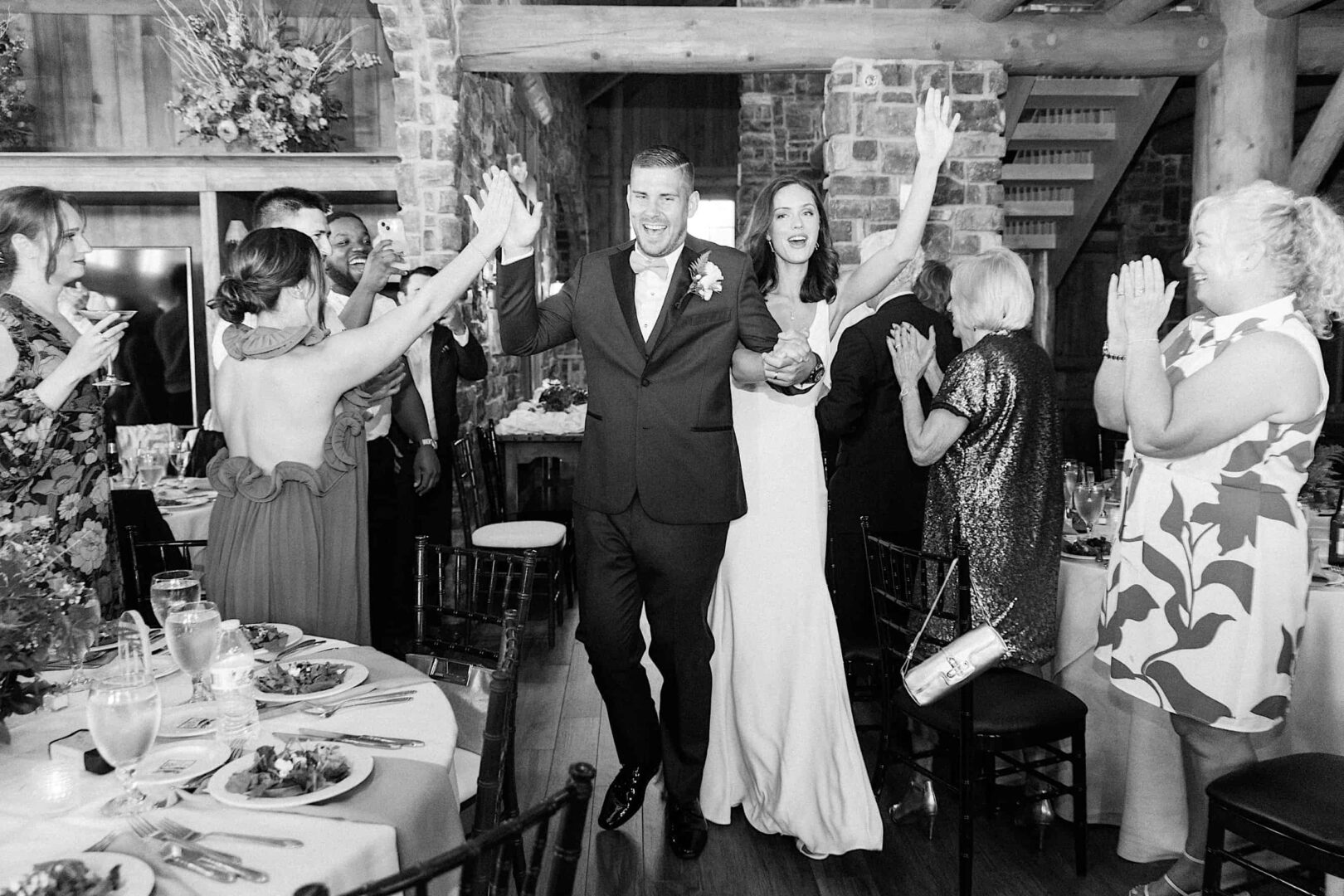 A newlywed couple in wedding attire walk into The Ridge at Back Brook reception hall in New Jersey, holding hands and smiling, as guests stand around clapping and celebrating. Tables are set with food and drinks.