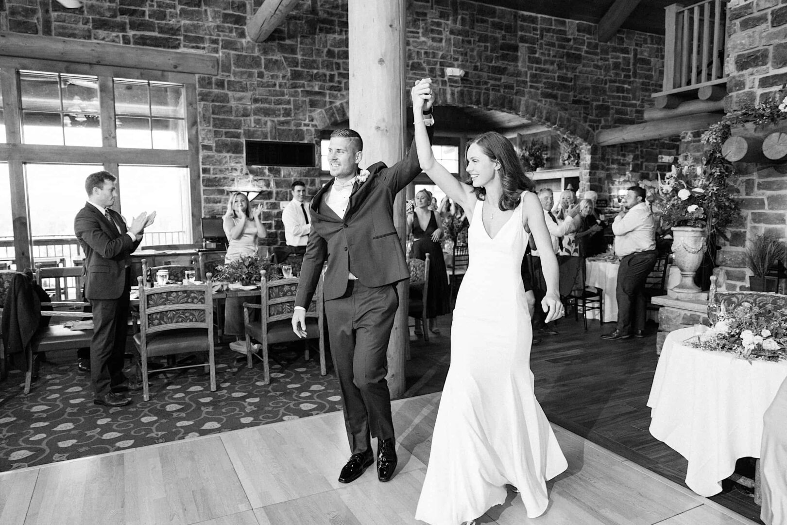 A newlywed couple walks into a reception hall at The Ridge at Back Brook, holding hands up in celebration as guests applaud. The setting features stone walls and wooden beams, creating an enchanting backdrop for their New Jersey wedding.