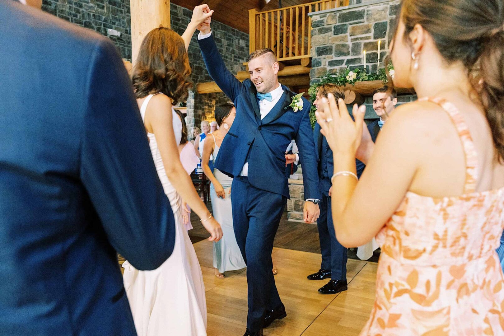 A man in a blue suit raises his arm while dancing at a wedding at a New Jersey country club, with other guests surrounding him and clapping.