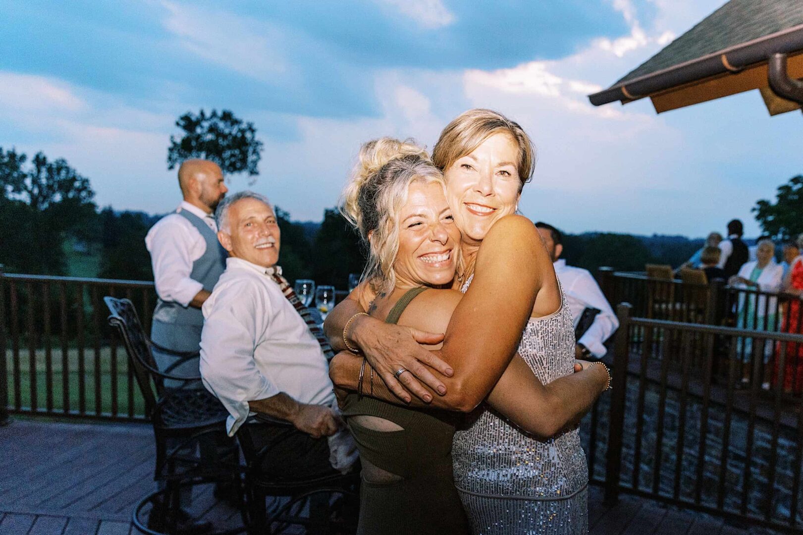 Two women embrace and smile for the camera on a deck at a wedding in New Jersey. A man seated at a table in the background also smiles. Other attendees are visible further back, enjoying the event at the elegant country club.