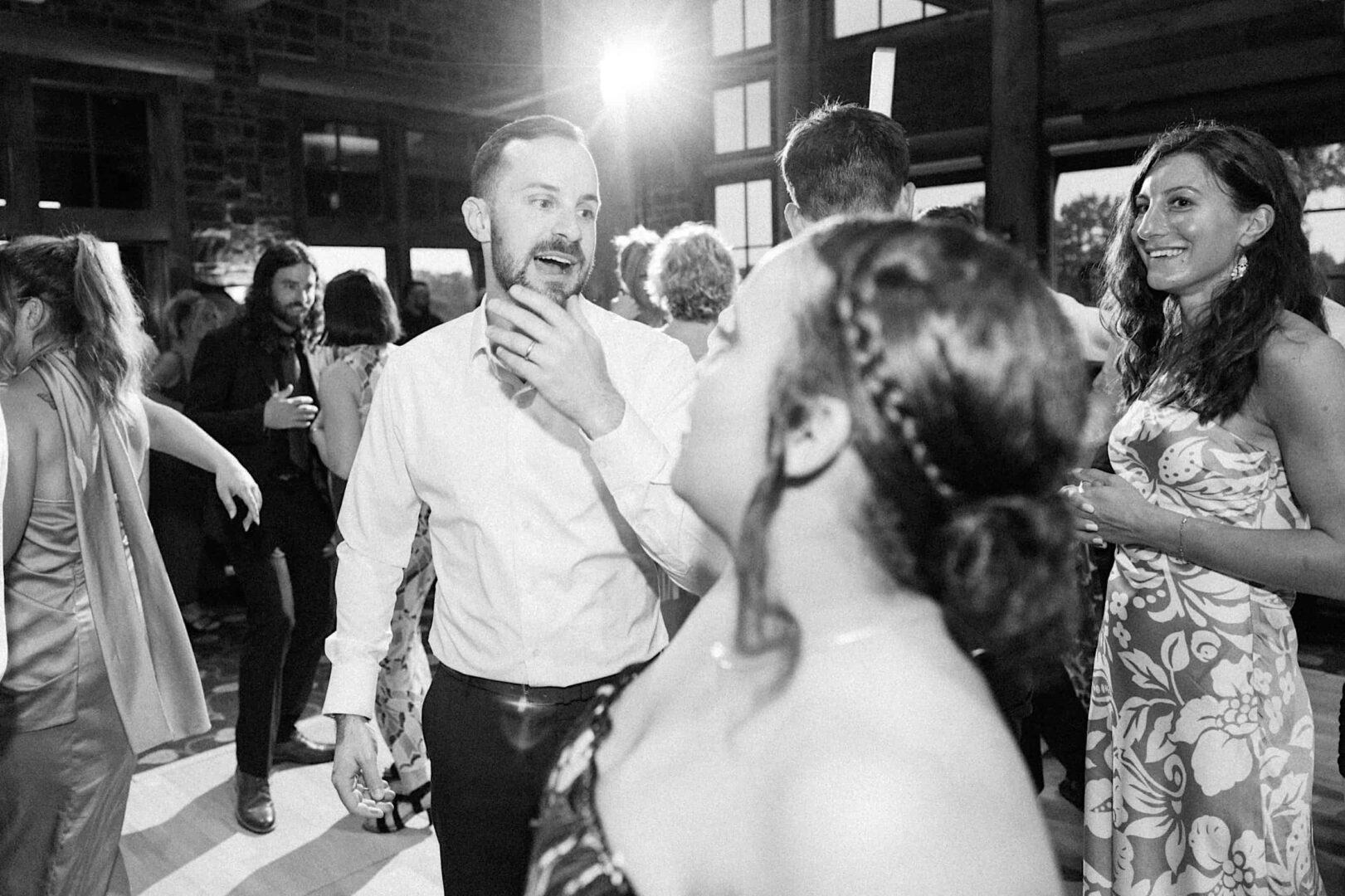 A black and white photo of people dancing at a wedding held at a country club in New Jersey. One man in the center is engaged in conversation while others dance around him. The background shows windows and decorative stone and wood details.