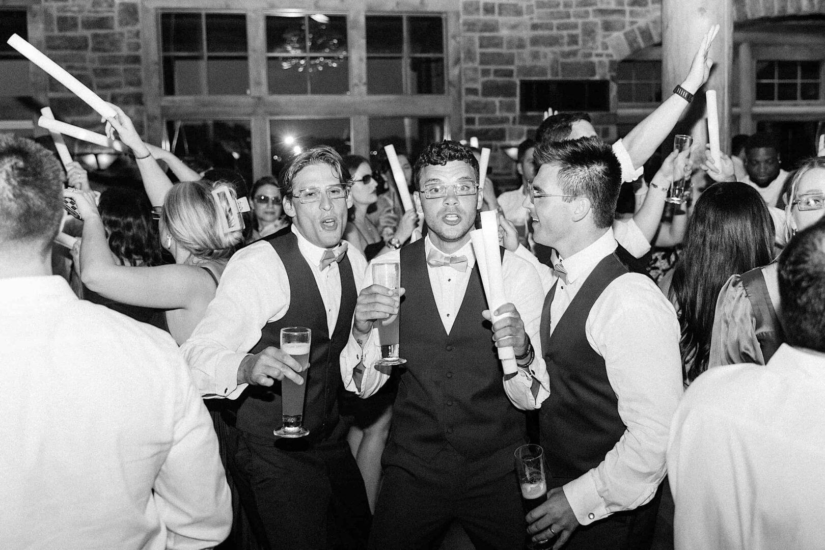 Black and white photo of a large group of people on a dance floor, some holding glowing sticks. At The Ridge at Back Brook in New Jersey, three men in suits are in the foreground, singing and holding drinks while others cheer behind them at what appears to be a lively wedding celebration.