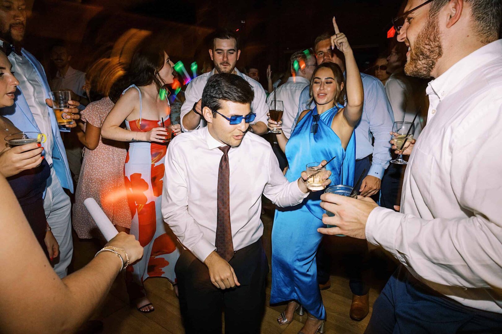 At The Ridge at Back Brook, a group of people in formal attire are dancing at an indoor party. One person in the center is wearing blue sunglasses, while others around them are holding drinks and engaging in lively conversations. It feels like a glamorous country club wedding celebration.