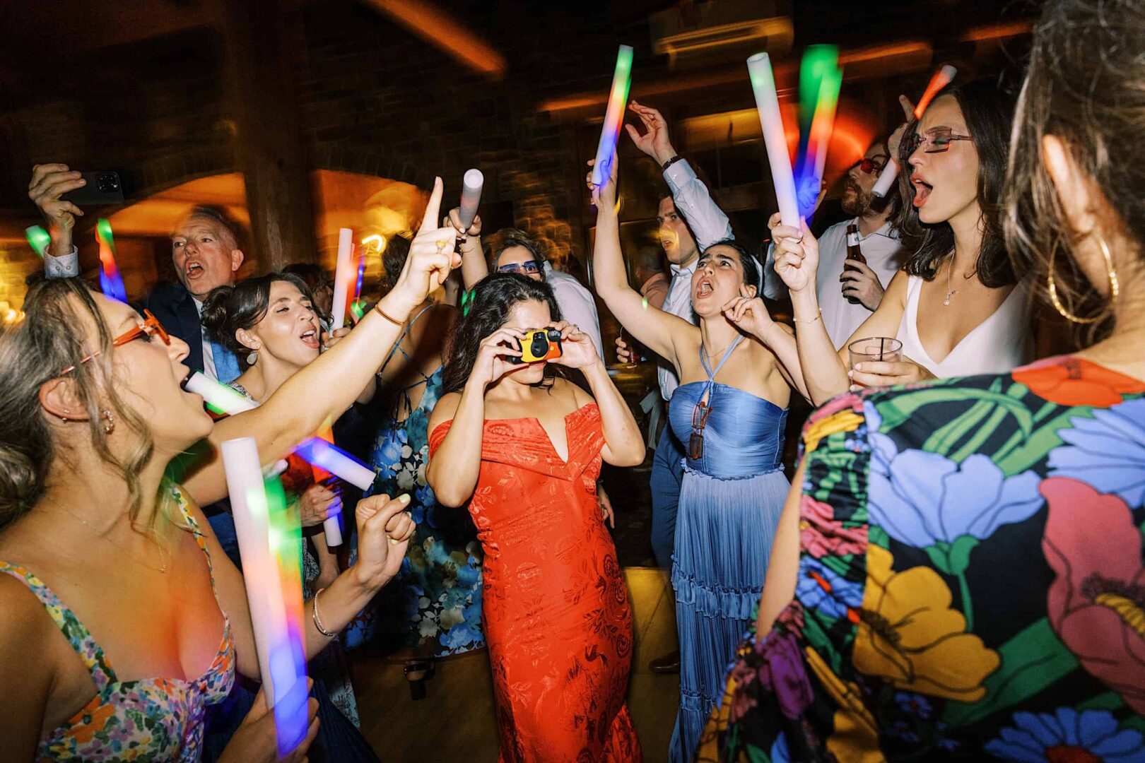 At The Ridge at Back Brook in New Jersey, a group of people dressed in formal wear are dancing with colorful glow sticks. One person in a red dress is taking a photo, while others around her appear to be singing and celebrating at the country club event.