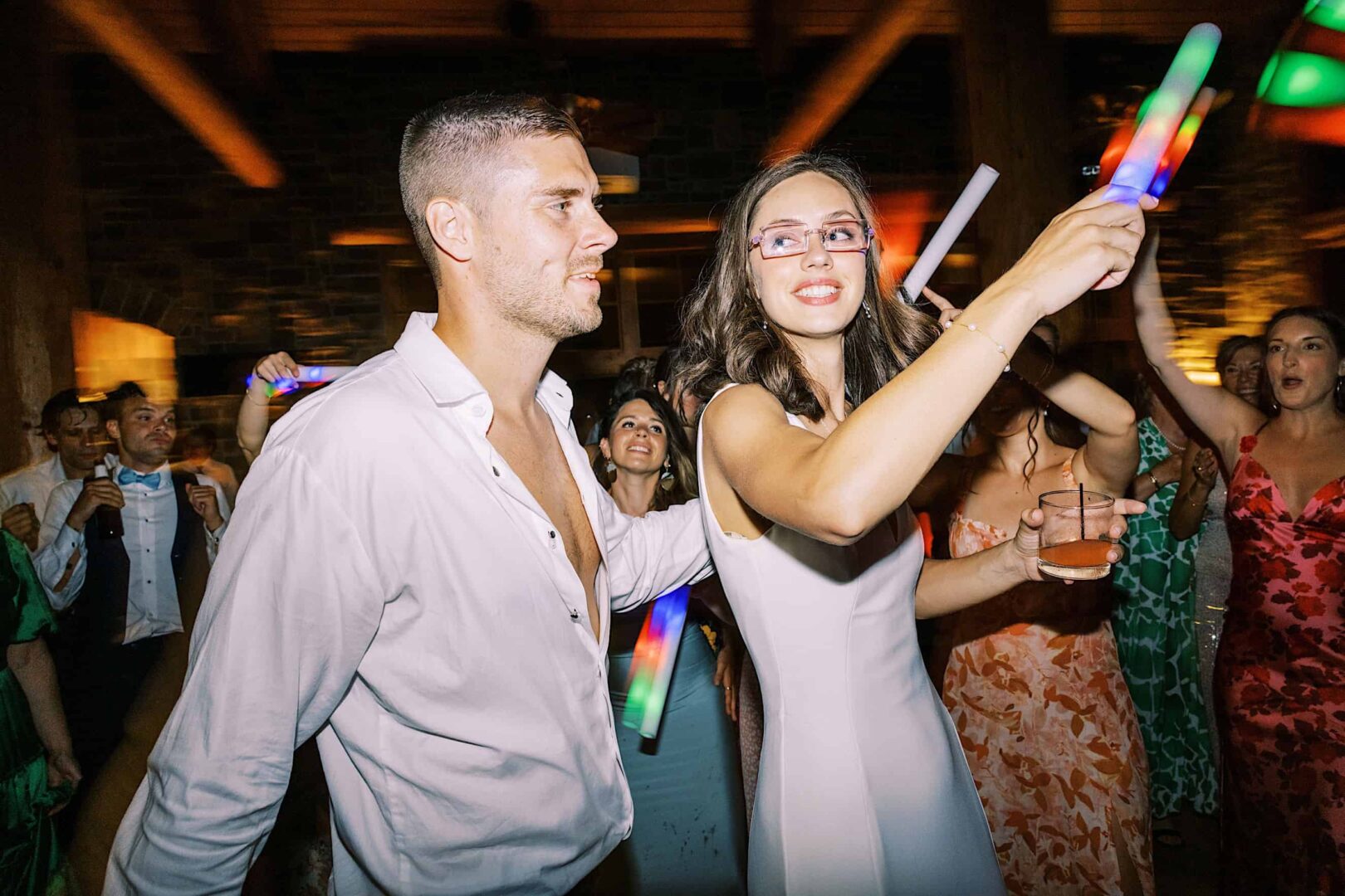 At a lively wedding reception in a New Jersey country club, a man and woman dance energetically in the crowd, holding colorful light sticks. The woman, with glasses and a drink in one hand, moves to the beat as dim lighting casts shadows over other joyful dancers in the background.