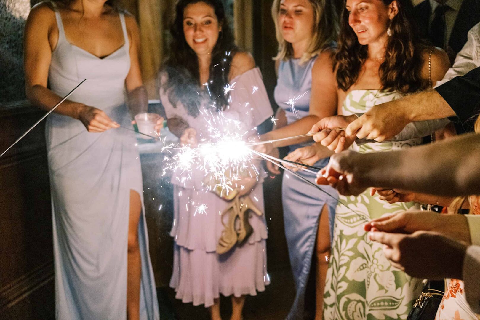 A group of people in semi-formal attire is holding and lighting sparklers indoors, as if capturing the magic of a charming wedding at a prestigious New Jersey country club.